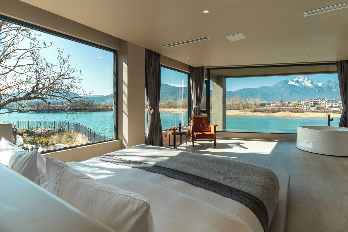 Ein Schlafzimmer mit einem großen Bett, einem Sessel, einem runden Tisch und Blick auf einen See und Berge.