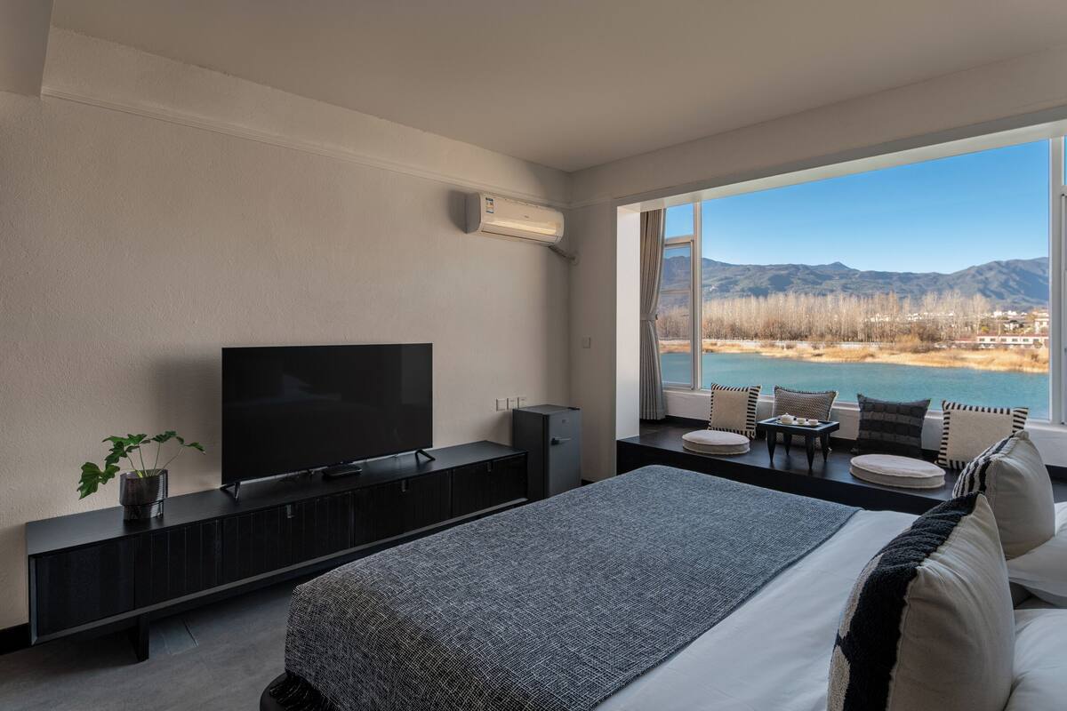 Ein modernes Schlafzimmer mit einem großen Bett, einem Flachbildfernseher, einem kleinen Tisch und Blick auf einen See und Berge.