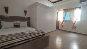 Une chambre d’hôtel avec un grand lit, une tête de lit, une fenêtre avec des rideaux et un sol carrelé.