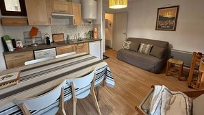 A modern kitchen with wooden cabinets, a dining area with a striped tablecloth, and a living room with a sofa and cushions.