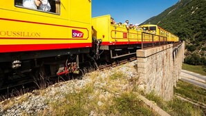 A yellow train with passengers, passing over a stone bridge.