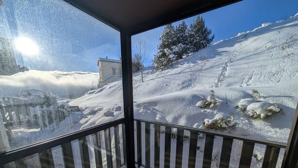 A snow-covered balcony with a clear view of a snowy landscape, including a tree and a building.