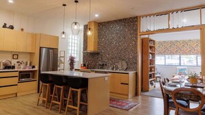Una cocina moderna con armarios de madera, una isla central con taburetes, iluminación colgante y vistas hacia un comedor con pared decorativa.
