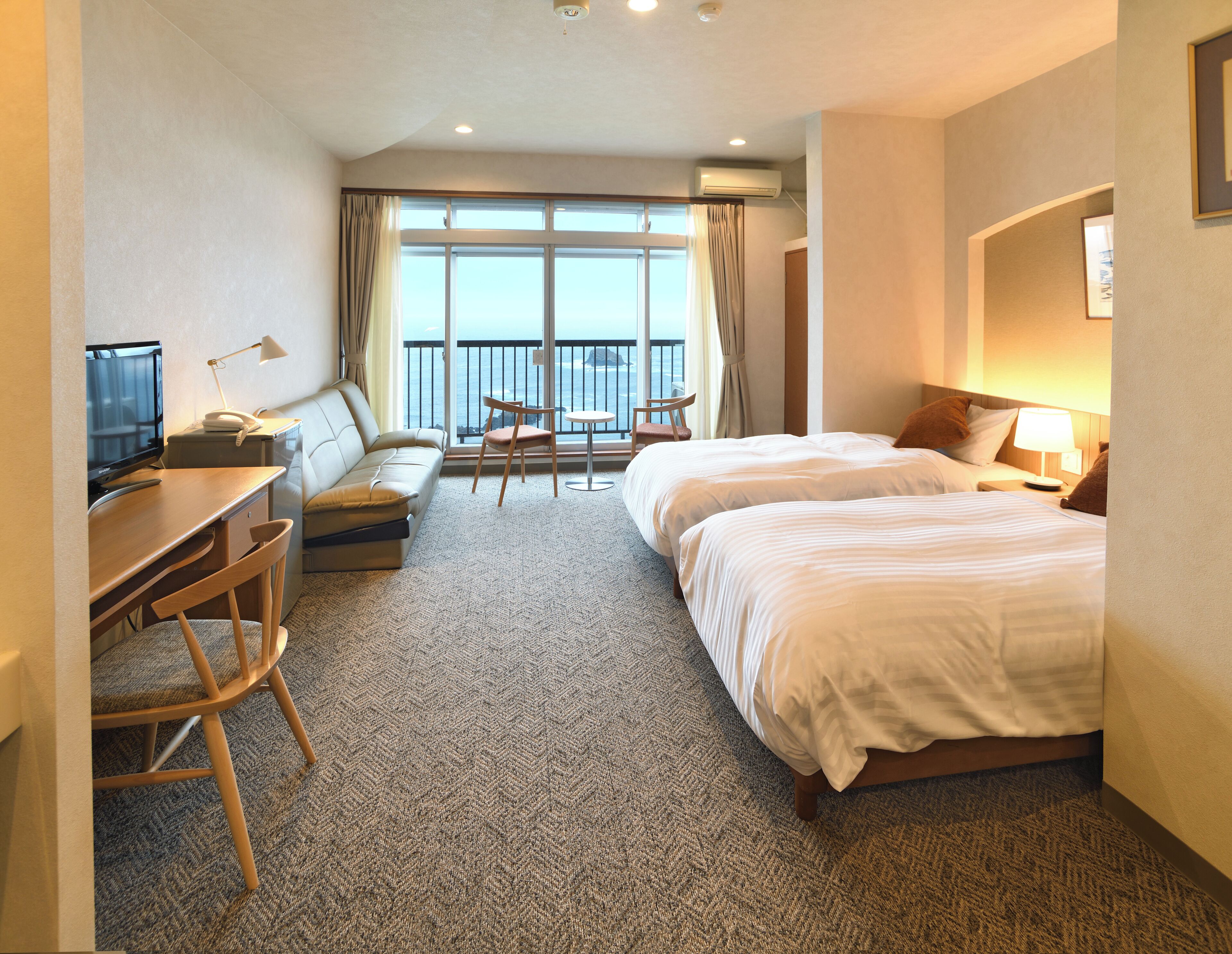 A hotel room with a desk, two beds, a sofa, a small table, and a balcony with a view.