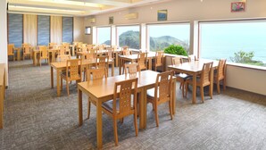 A spacious dining area with multiple wooden tables and chairs, large windows offering a view of the sea and hills, and a carpeted floor. - SADO Futatsugame View Hotel (Sado)