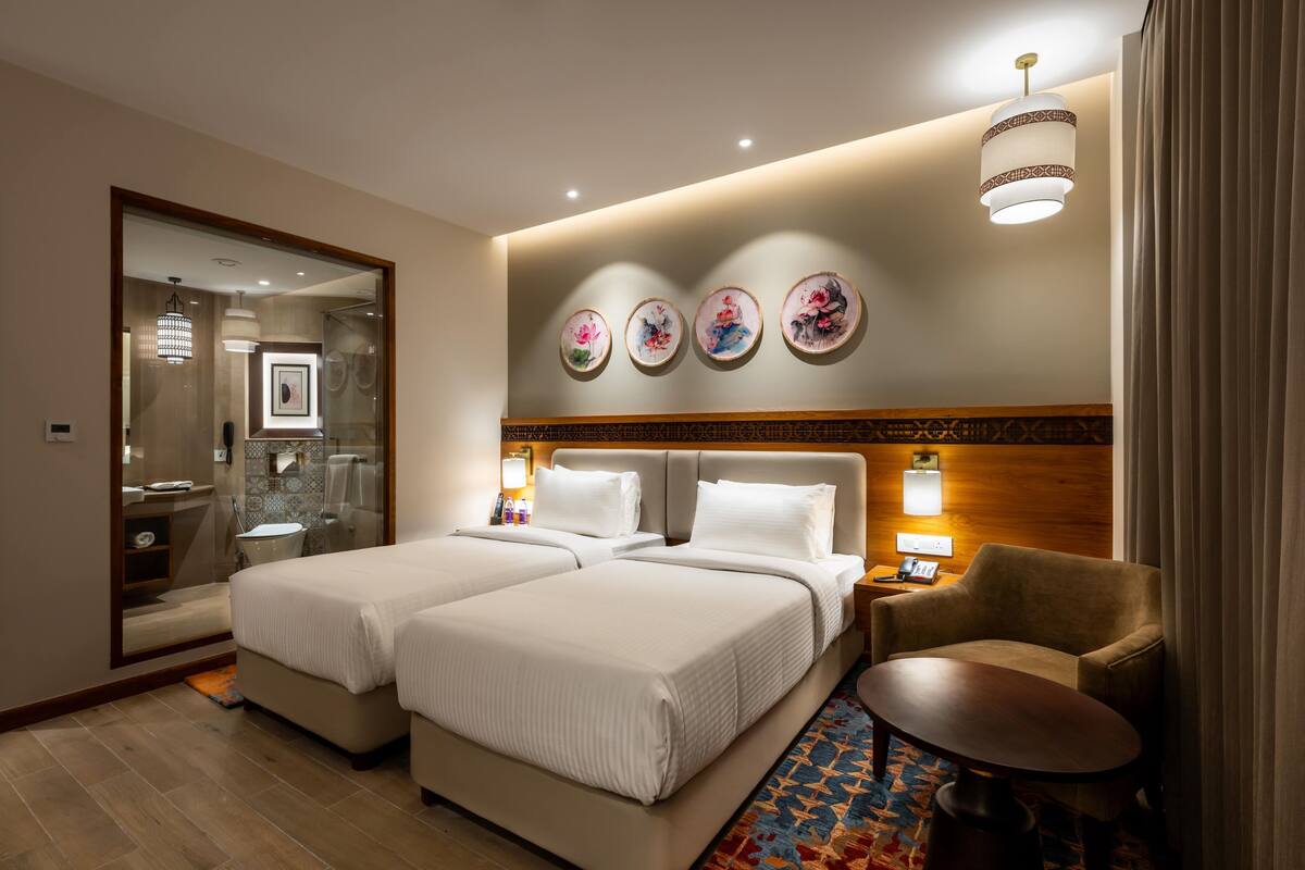A hotel room with two beds, a sitting area with a chair, a small table, a mirror, and a bathroom visible through the glass door.