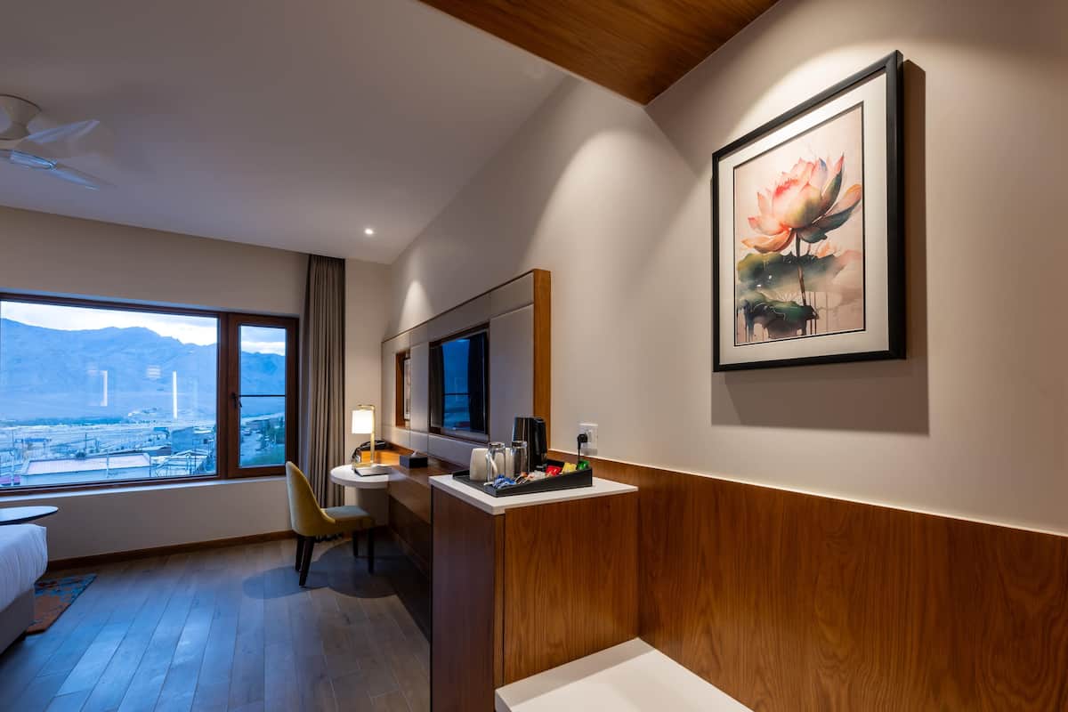 A hotel room with a bed, a desk with a chair, a window with a view of mountains, and a framed artwork on the wall.