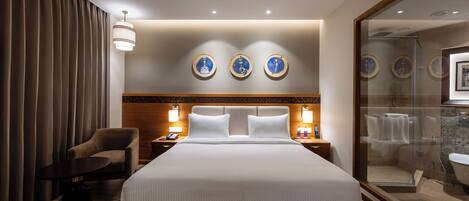 A hotel room with a large bed, a chair, a bedside table, a nightstand, and a glass-enclosed shower area.