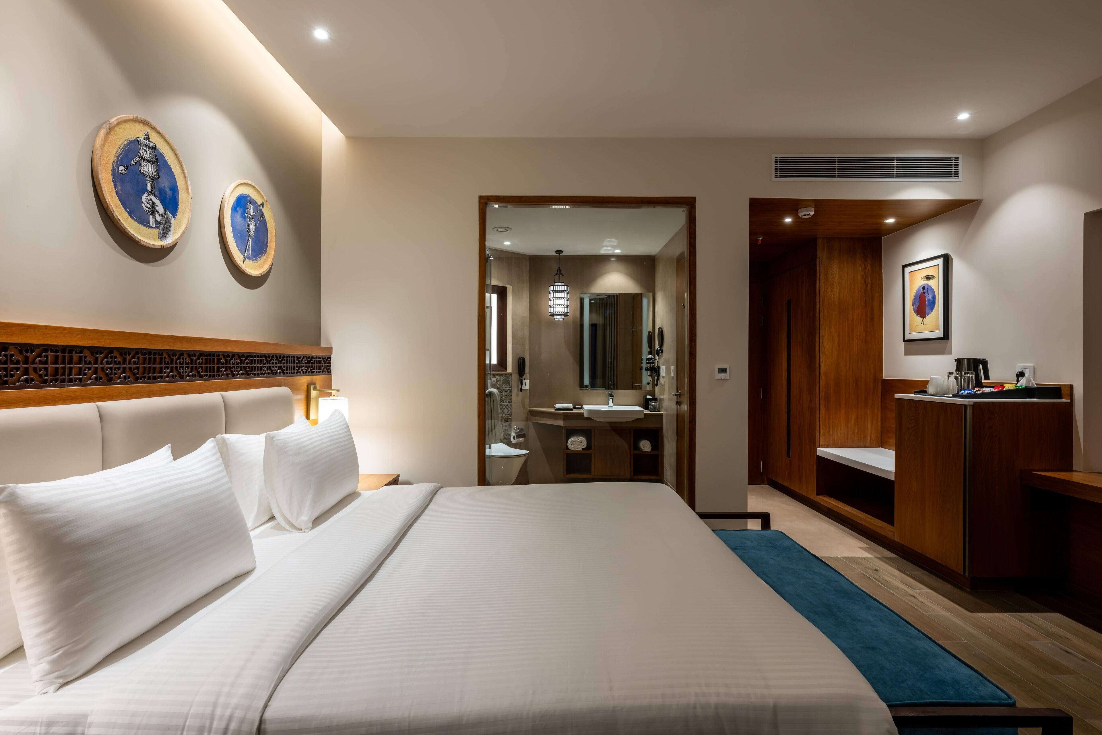 A hotel room with a large bed, a wooden headboard, a bedside table, a mirror, a wooden wardrobe, and a bathroom with a sink and mirror.