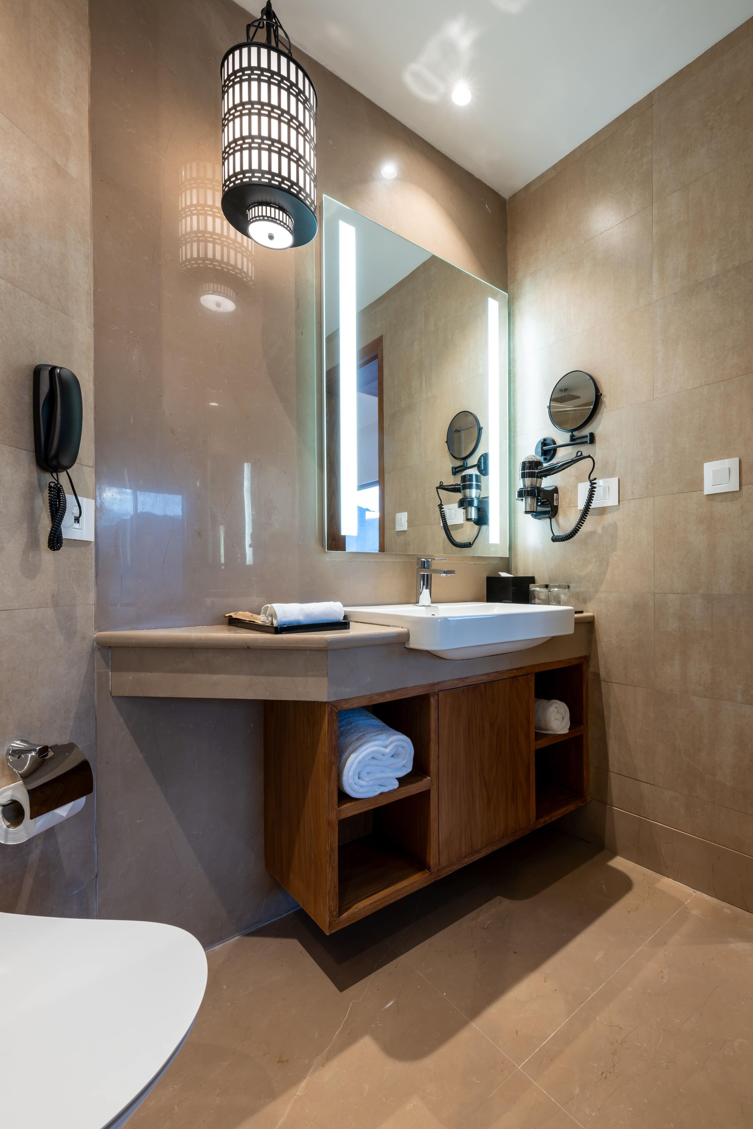 A modern bathroom with a large mirror, a sink, and a toilet. There is a wooden vanity with shelves, a hairdryer, and a phone on the wall.