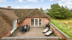 Ein Haus mit Strohdach, einer Ziegelterrasse, einem Esstisch mit Stühlen und zwei Liegestühlen.