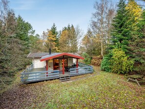 Une cabane avec un toit rouge et une terrasse, entourée d’arbres et d’une zone herbeuse.