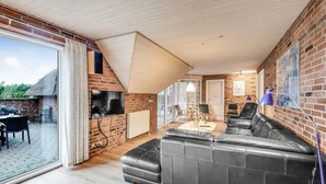 En moderne stue med en sort lædersofa, et fjernsyn monteret på en murstensvæg og en stor skydedør til en udendørs terrasse.