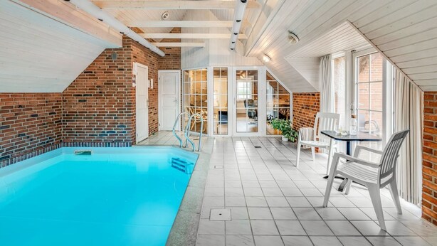 En indendørs swimmingpool med flisegulv, murstensvæg og loft med træbjælker.