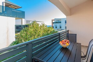 A balcony with a table and chairs, a bowl of fruit, and a view of buildings and greenery. - Apartments Markovic (Vir)
