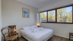 A bedroom with a bed, a chair, a bedside table, a lamp, a picture on the wall, and a window with a view of trees and cars. - The Balcony of Sainte-Anne, Lake View (Annecy-le-Vieux)