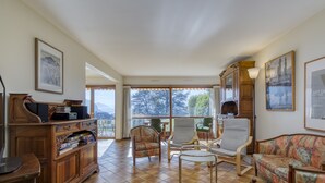 A spacious living room with a large window, wooden furniture, and framed artwork. - The Balcony of Sainte-Anne, Lake View (Annecy-le-Vieux)