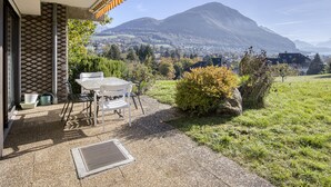 A patio with a table and chairs, a view of a mountain, and a building with a patterned wall. - The Balcony of Sainte-Anne, Lake View (Annecy-le-Vieux)