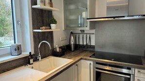 Una cucina moderna con lavello in acciaio inossidabile, forno integrato e cappa aspirante. Sopra il lavello si trovano scaffali in legno e una finestra a sinistra.
