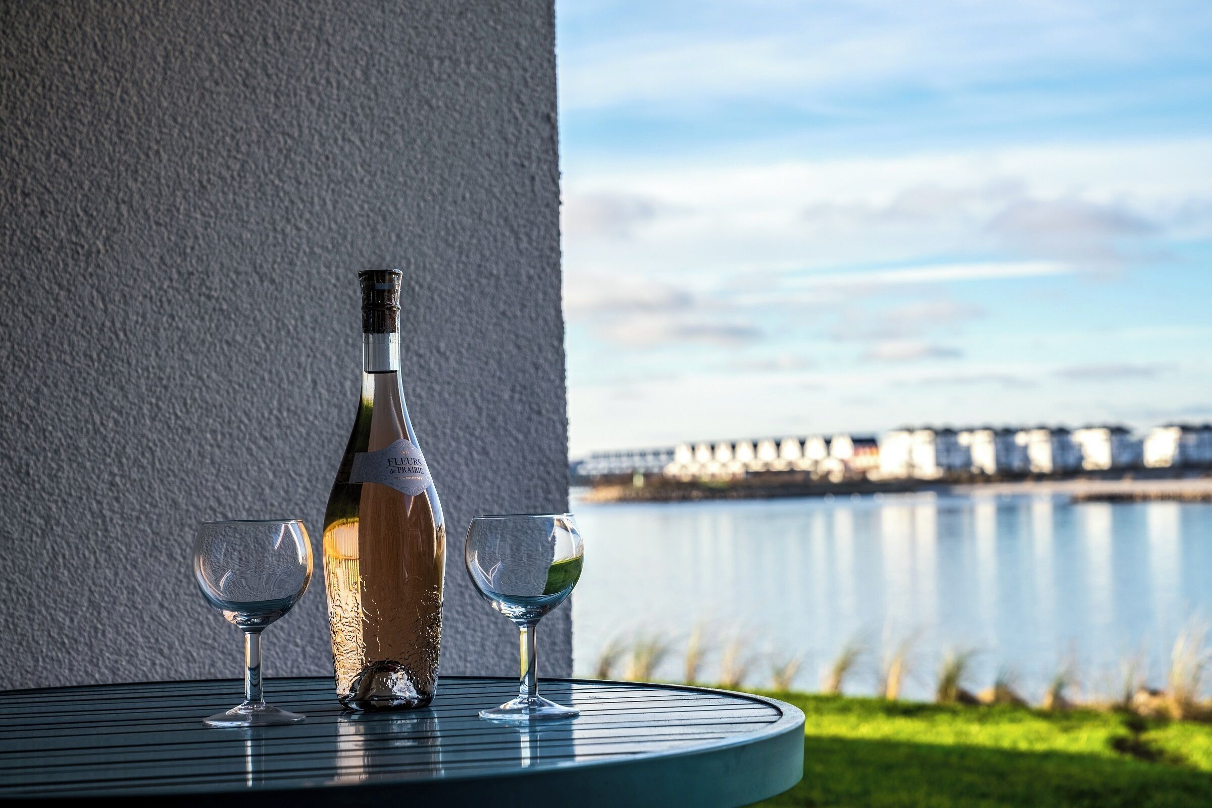 Una bottiglia di vino e due bicchieri su un tavolo rotondo, con vista su un corpo d'acqua e un ponte sullo sfondo.