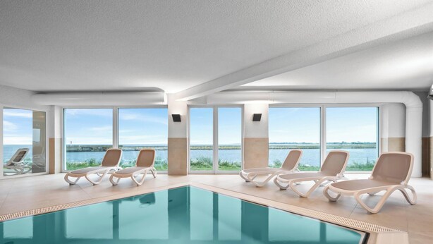Piscina interior moderna con vistas claras al agua exterior, rodeada de sillones de descanso.