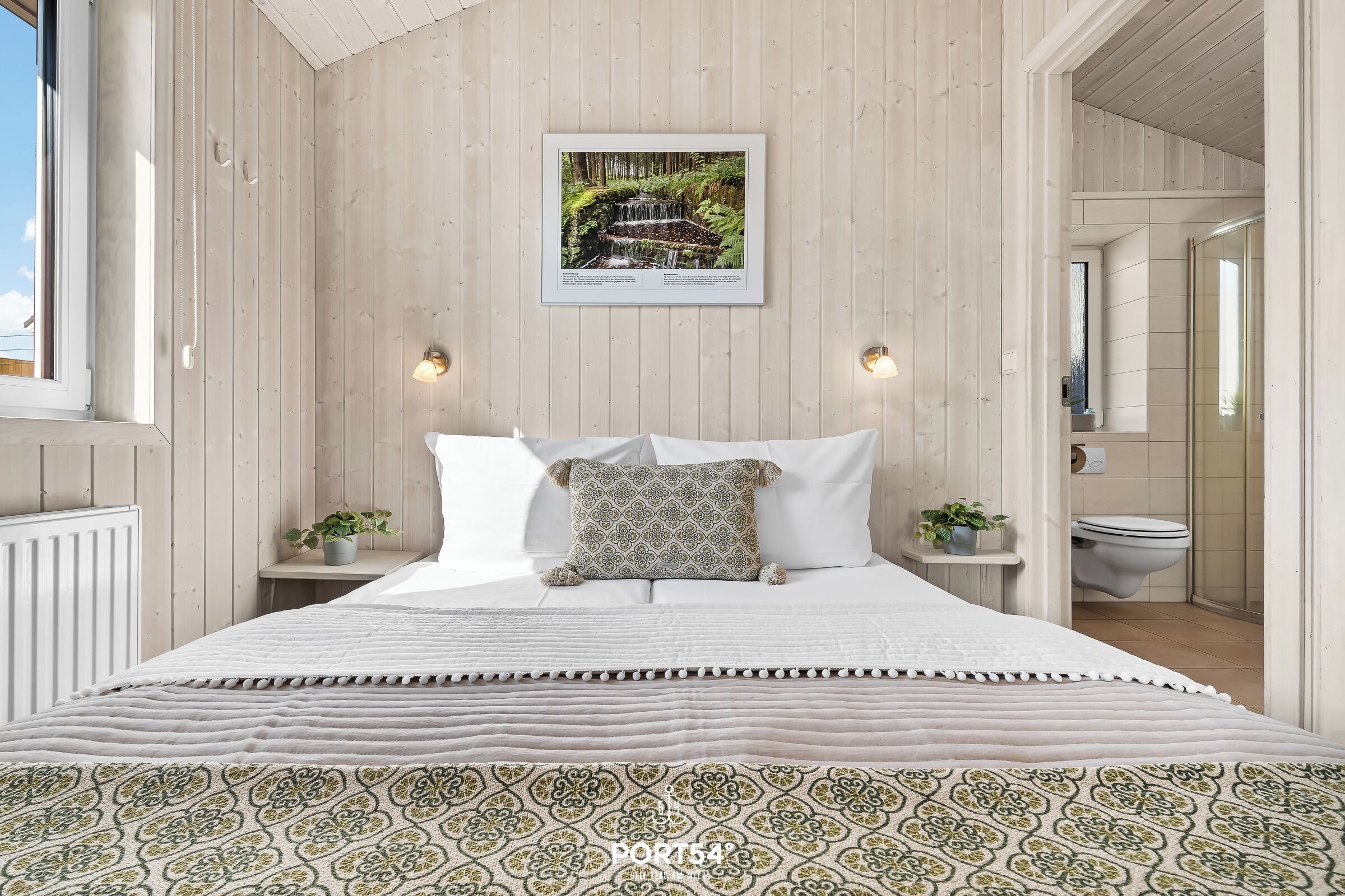 Ein Schlafzimmer mit einem Bett, einem gerahmten Bild an der Wand, Nachttischen mit Lampen und einem Fenster.