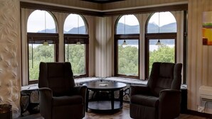A room with two leather chairs, a round table, and large arched windows offering a view of mountains and trees. - Hulder- og Trollparken (Senja)
