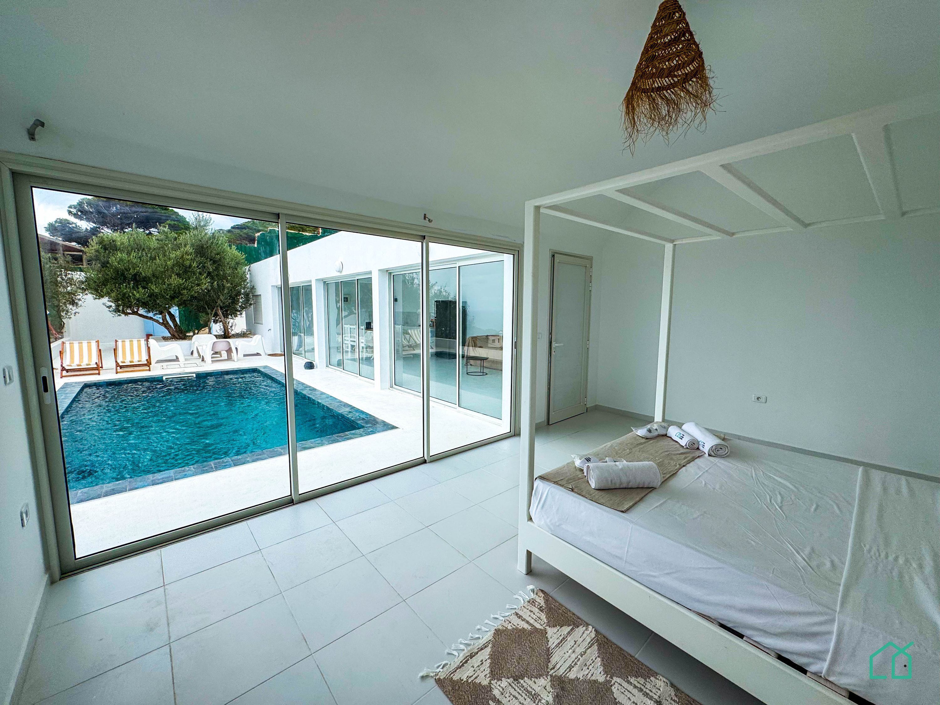 Un dormitorio moderno con un amplio ventanal que da a una piscina y zona de estar exterior.