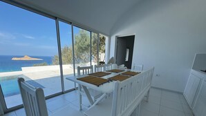 Un comedor con una mesa y sillas blancas, ventanas amplias con vistas al mar y a la playa, y suelo de baldosas.
