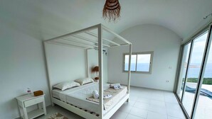 Un dormitorio con cama con dosel, una mesita de noche pequeña, una mesita auxiliar, una alfombra estampada y un ventanal con vistas al mar.