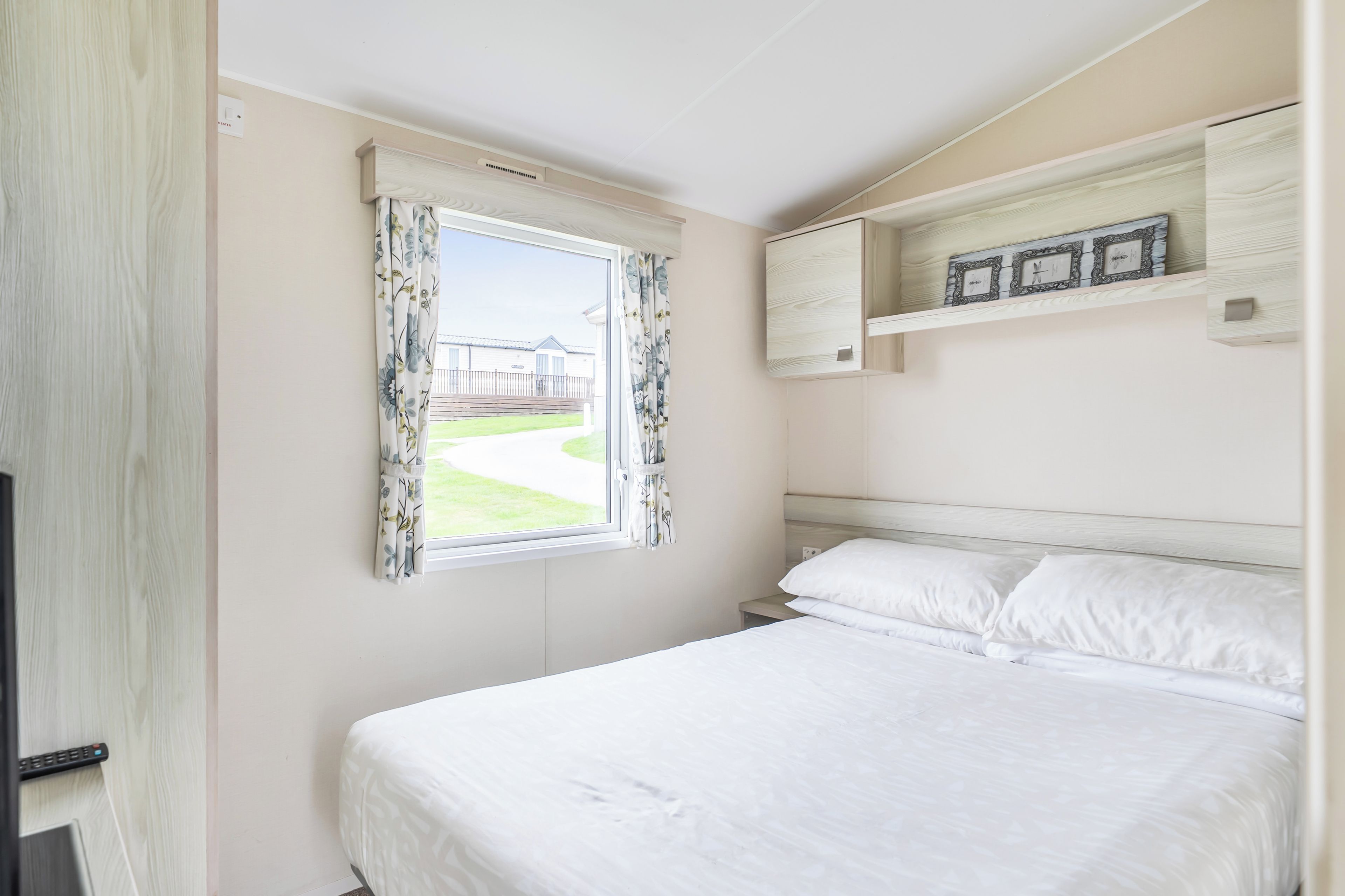 Une chambre avec un lit simple, une fenêtre ornée de rideaux à motifs floraux, une étagère intégrée avec des cadres de photos, et une vue sur un bâtiment à l’extérieur.