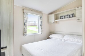Un dormitorio con una cama individual, una ventana con cortinas florales, una estantería empotrada con marcos de cuadros y vistas a un edificio.