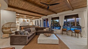 A spacious living area with a large sectional sofa, a coffee table, and a dining area with chairs. A ceiling fan is visible, and there are large windows offering a view of the outside. - Luxe Villa Tulum in Indio (Indio)