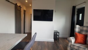 Una sala moderna con un televisor de pantalla plana grande montado en la pared, un sofá a cuadros con un cojín naranja, una mesa de comedor con sillas y una pequeña mesita auxiliar negra.