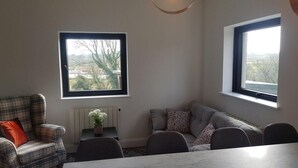 Sala de estar con una mesa de comedor, dos sofás y dos ventanas que dan a un árbol y un paisaje lejano.