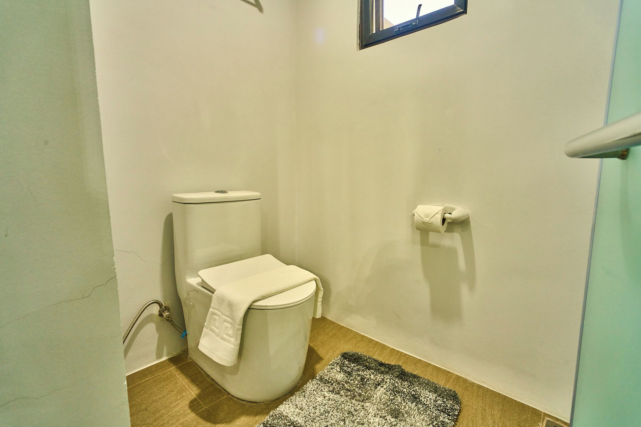 Un baño pequeño y funcional con inodoro, una toalla, un tapete y una ventana.