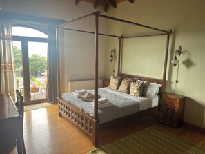 A four-poster bed with a wooden frame, a bedside table with a lamp, and a view of the outdoors through a glass door.