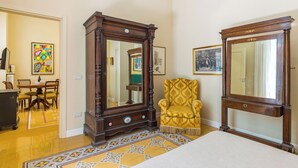 A room with a large wooden dresser, a yellow armchair, and a bed. There is a dining table with chairs and a colorful rug. - Santa Lucia Seaview (Naples)