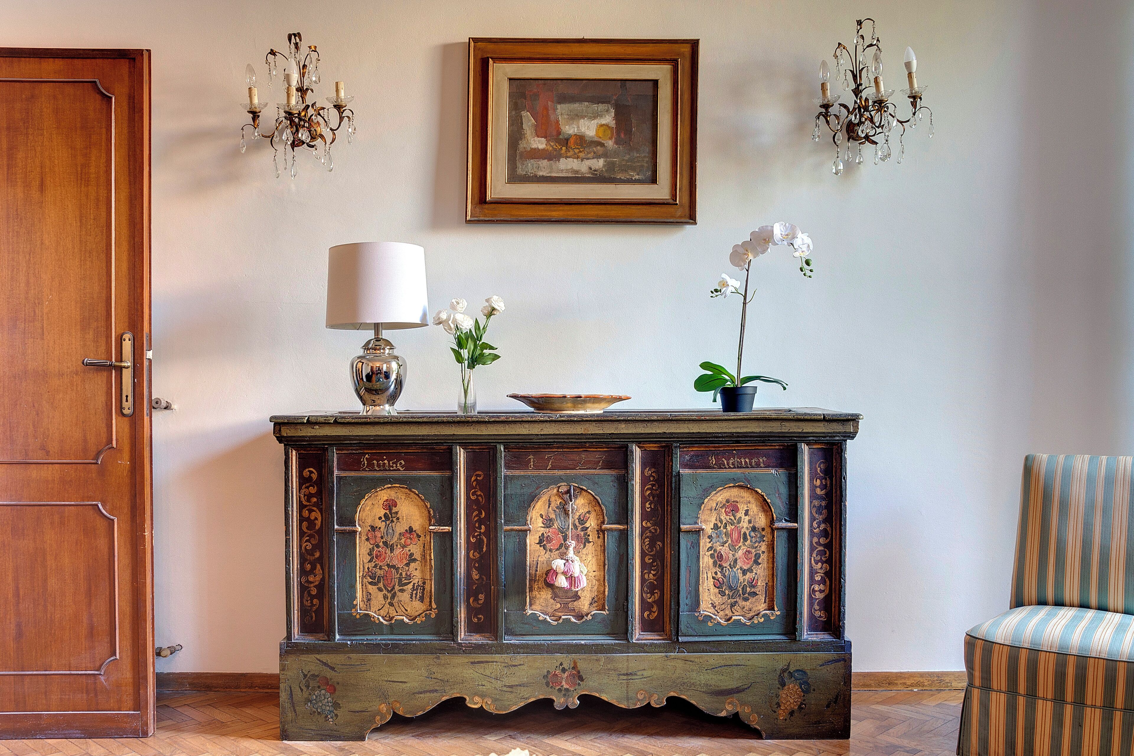 Una puerta de madera, un aparador con diseños florales, un cuadro enmarco y una araña.
