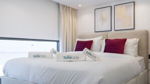 Ein Hotelzimmer mit einem großen Bett, weißer Bettwäsche und zwei roten Kissen. An der Wand hängen zwei gerahmte Kunstwerke und ein Fenster mit Vorhängen.