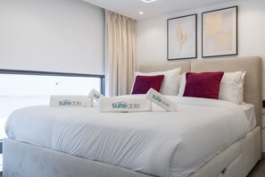 A hotel room with a large bed, white bedding, and two red pillows. There are two framed artworks on the wall and a window with curtains. - Trendy Loft Studio in Oxford Terrace JVC (Dubai)