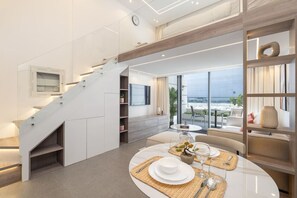 A modern hotel room with a dining area, a staircase, and a large window offering a view of the beach.