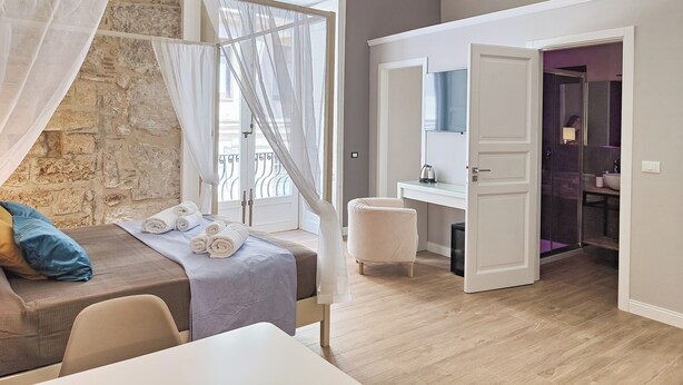A bedroom with a stone wall, a bed with rolled towels, a bedside table, and a bathroom with a mirror and sink. - Dimora Carella (Palermo)
