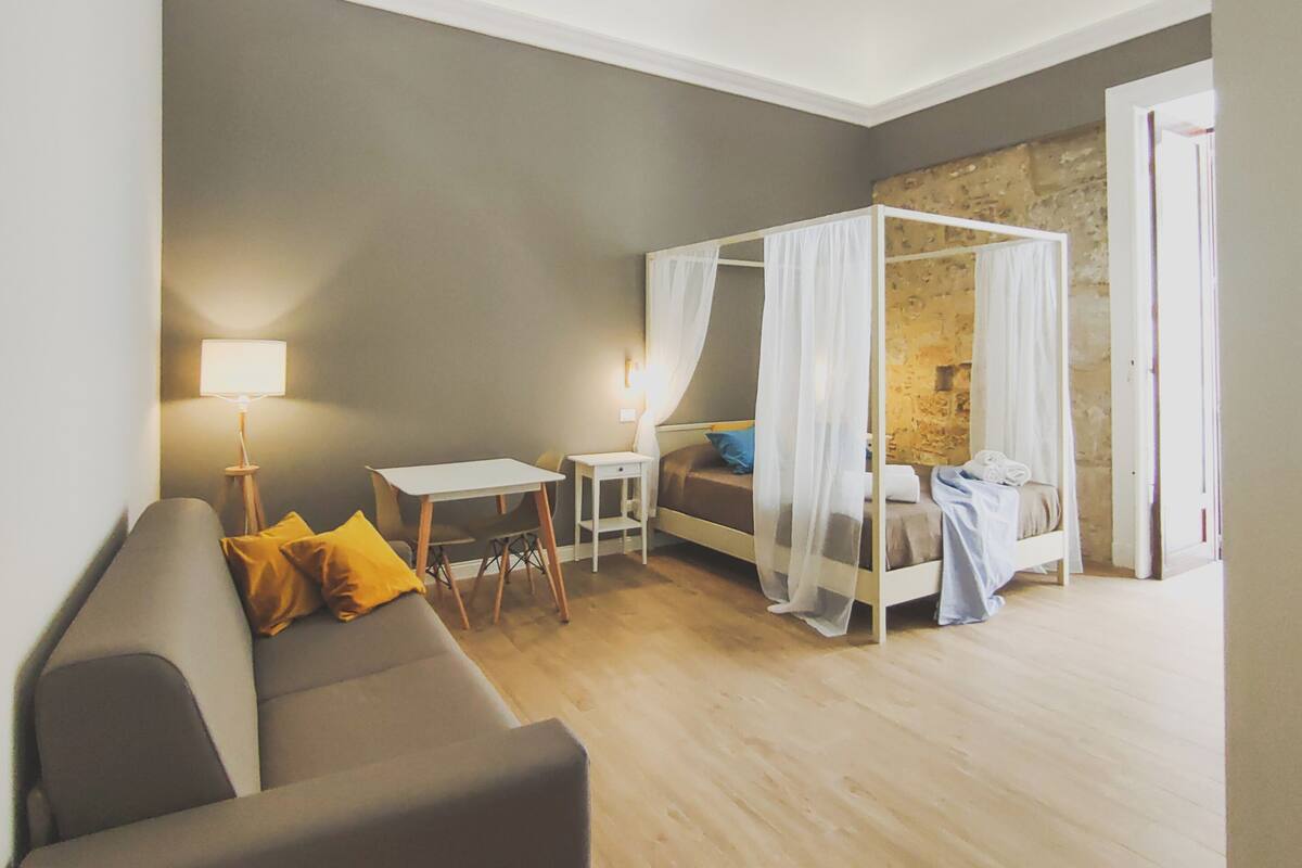 Quarto moderno com sofá cinza, uma mesinha, mesa de cabeceira, cama com dossel e parede de pedra.