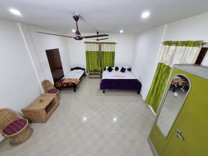 A compact hotel room with two single beds, a ceiling fan, a small table, a wicker chair, and a green cabinet.