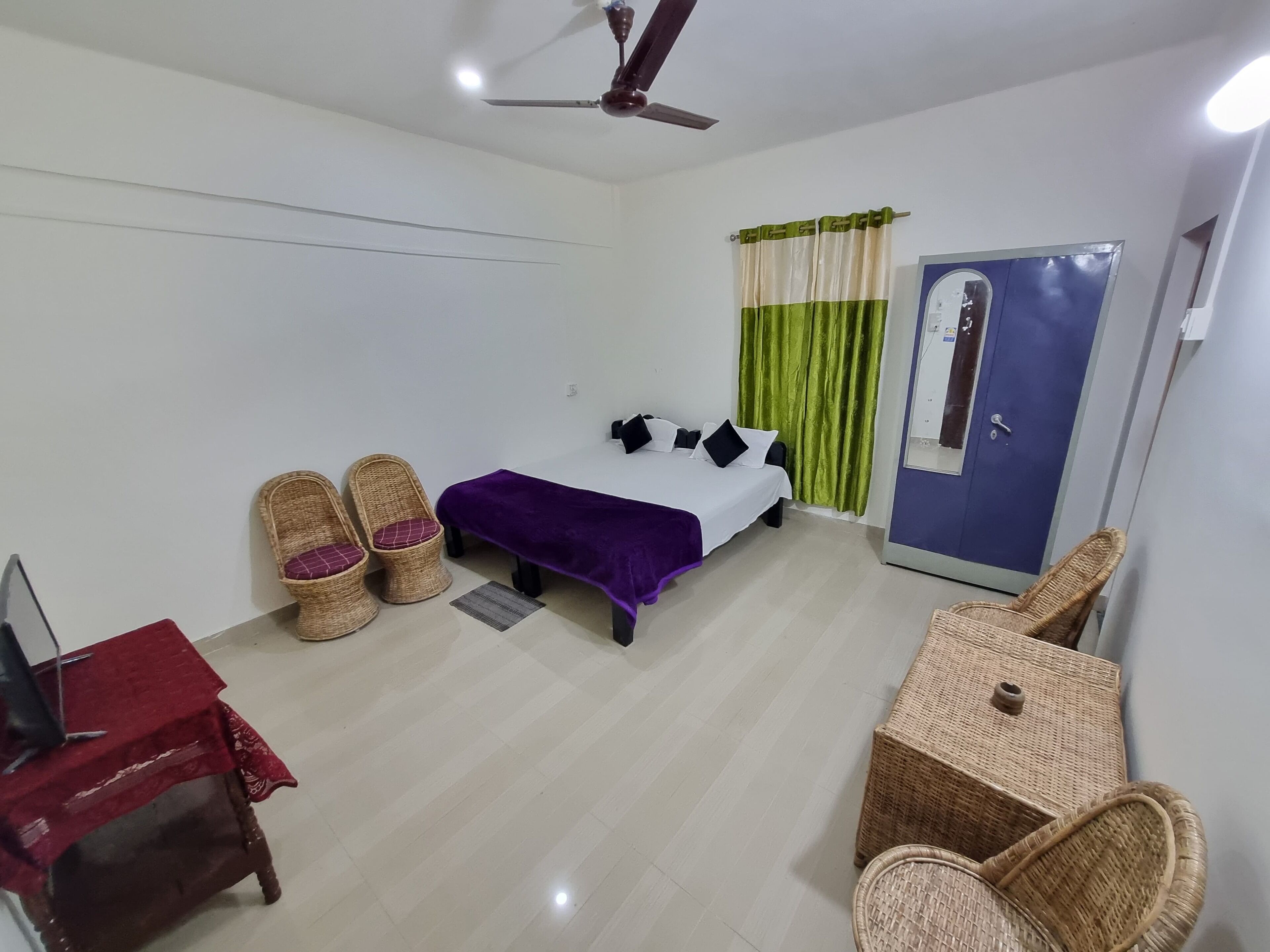 A room with a bed, a desk, two wicker chairs, a ceiling fan, and a door with a mirror.