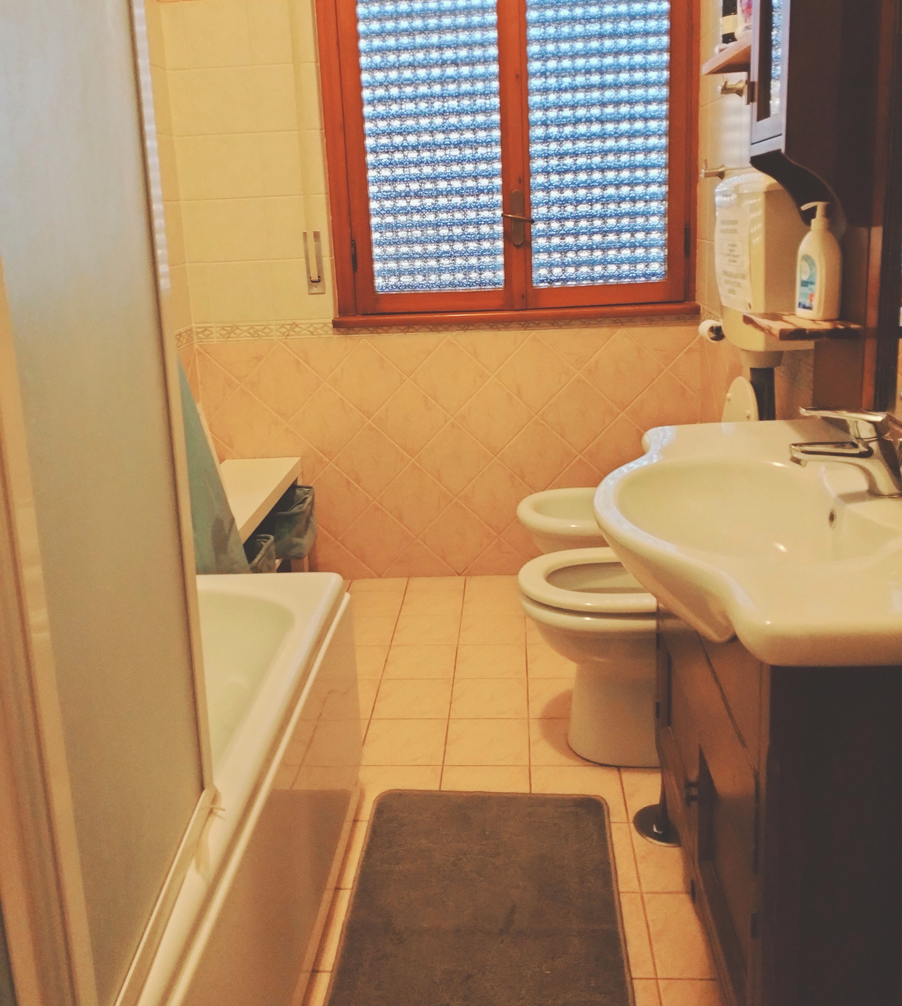 Une petite salle de bain avec des toilettes, un lavabo et une baignoire. Il y a une fenêtre avec un rideau à motifs.