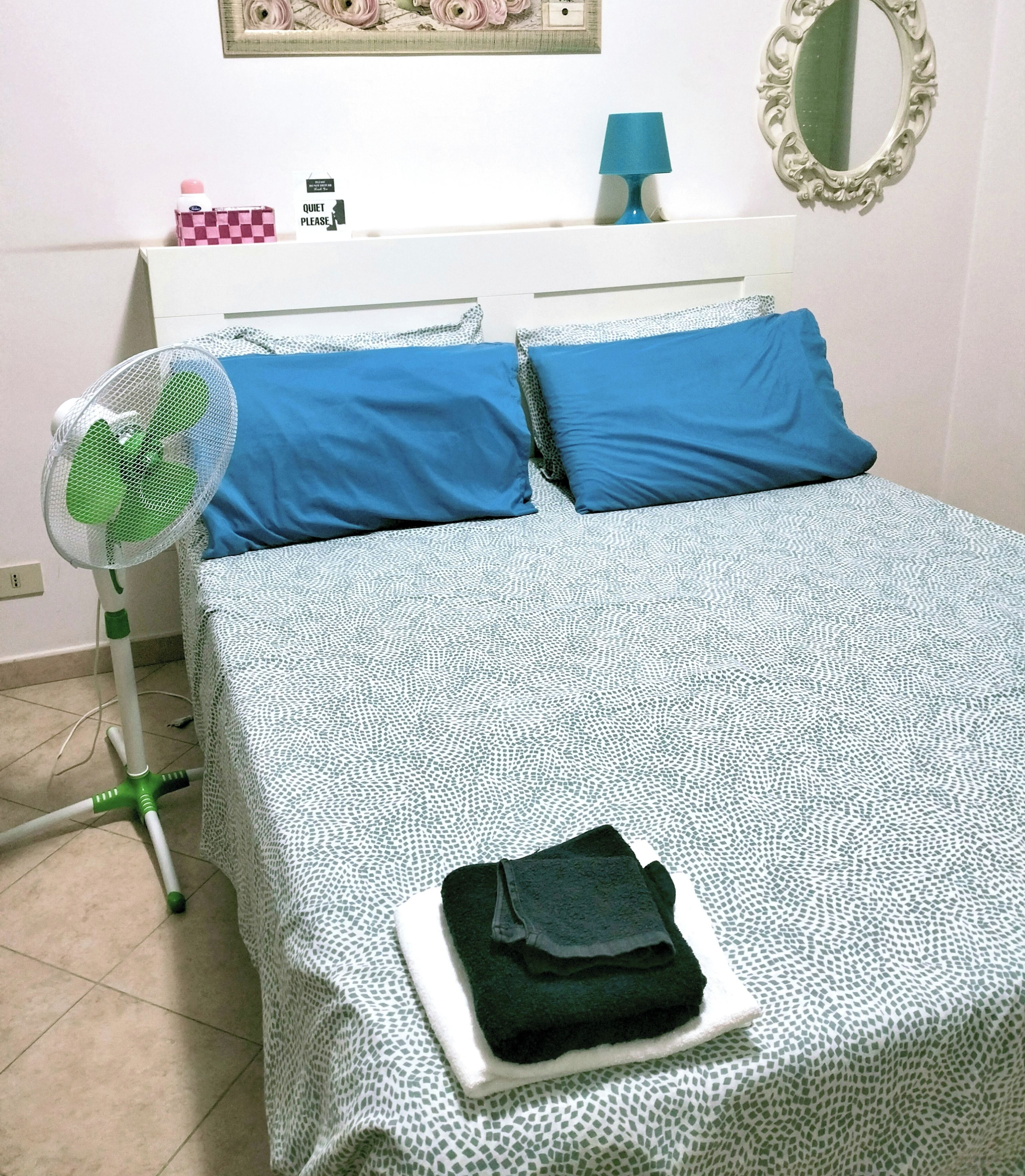 A neatly made bed with blue pillows and a white patterned bedspread, a standing fan, a small table with a lamp, and a mirror on the wall.