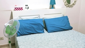 Ein ordentlich gemachtes Bett mit blauen Kissen und einer weißen, gemusterten Bettdecke, ein Standventilator, ein kleiner Tisch mit Lampe und ein Spiegel an der Wand.