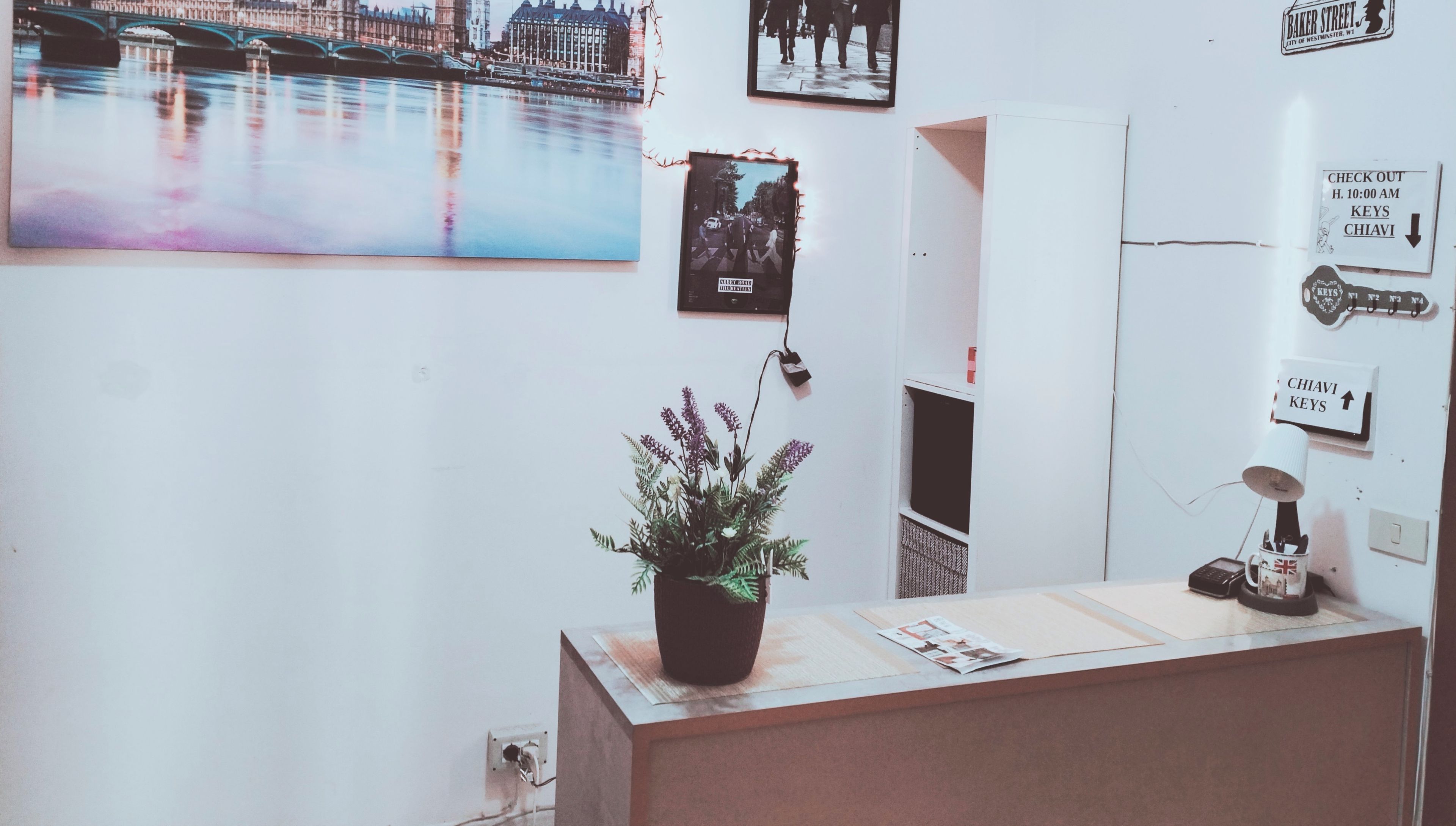 Un mostrador de recepción con una pintura de paisaje urbano, una planta en maceta y un panel con llaves en la pared.